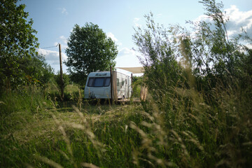 A camper van with a wooden terrace and fabric canopy is parked in the middle of tall grass and trees. This peaceful scene evokes a sense of freedom, glamping, and off-grid living.