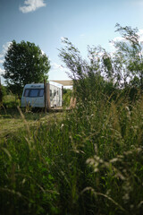 A camper van with a wooden terrace and fabric canopy is parked in the middle of tall grass and trees. This peaceful scene evokes a sense of freedom, glamping, and off-grid living.