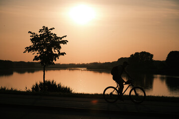 Silhouette of a man cycling past a lake at sunset, reflected in the water. Golden hour