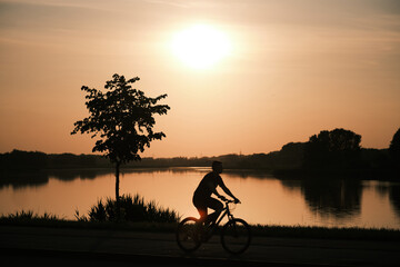 Silhouette of a man cycling past a lake at sunset, reflected in the water. Golden hour