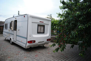 A white camper trailer is parked on a paved backyard near a brick house and cherry tree. RV travel content, camping promotions, suburban lifestyle visuals, or articles about alternative housing