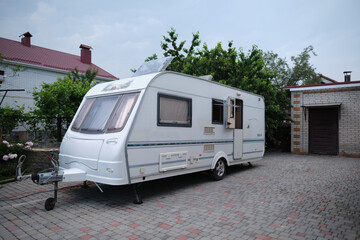 A white camper trailer is parked on a paved backyard near a brick house and cherry tree. RV travel content, camping promotions, suburban lifestyle visuals, or articles about alternative housing