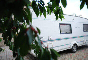 A white camper trailer is parked on a paved backyard near a brick house and cherry tree. RV travel content, camping promotions, suburban lifestyle visuals, or articles about alternative housing