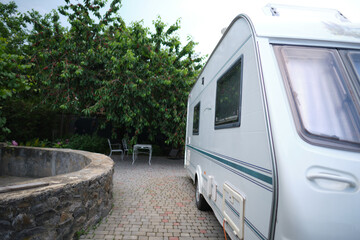 A white camper trailer is parked on a paved backyard near a brick house and cherry tree. RV travel content, camping promotions, suburban lifestyle visuals, or articles about alternative housing