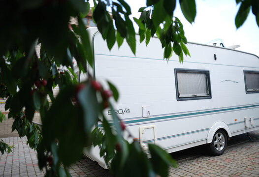 A white camper trailer is parked on a paved backyard near a brick house and cherry tree. RV travel content, camping promotions, suburban lifestyle visuals, or articles about alternative housing