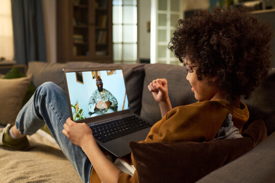 Black child lying on sofa holding video call with Black man in military uniform on laptop screen, showing family connection and communication with deployed parent at home