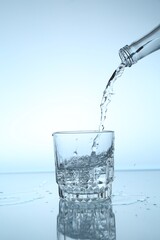 Pouring water from bottle into glass at mirror table against light blue gradient background, closeup