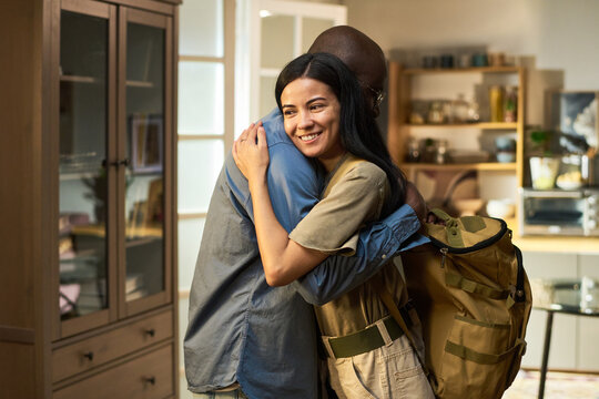 Young adult Black man in military uniform embracing young adult Caucasian woman indoors, both smiling, military backpack visible, showing reunion of military man and his family at home - Powered by Adobe