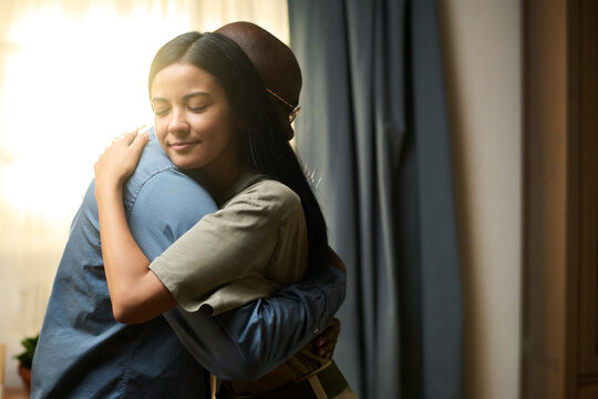 Young adult Asian woman embracing middle aged Black man in military uniform at home, both standing closely with eyes closed, showing emotional family reunion after deployment