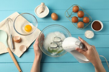 Woman whisking egg whites with hand mixer at light blue wooden table, top view
