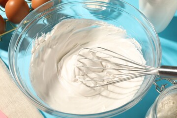 Whisk and whipped cream in bowl on light blue table, closeup