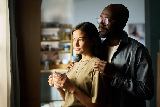 Caucasian young adult woman holding mug standing by window with Black middle aged man gently placing hands on her shoulders