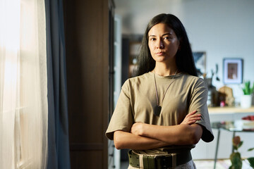 Portrait of young adult Asian woman standing with arms crossed wearing military dog tags at home,...