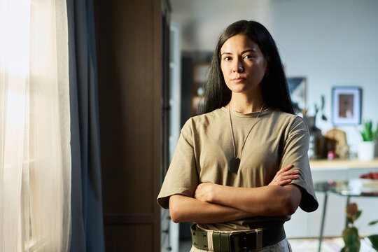 Portrait of young adult Asian woman standing with arms crossed wearing military dog tags at home, looking confidently at camera, background showing domestic interior details - Powered by Adobe