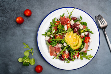 Green salad with avocado, salad leaves, jamon serrano or prosciutto, olives and tomatoes. Healthy diet lunch. Top view at dark blue stone table. High quality photo