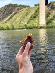 A female hand holding a broken piece of waffle cake (Andruty) in front of the Moselle River in Dieblich, Germany. Terraced vineyards and a bridge pillar in the background.