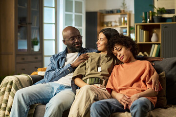 Black middle aged man in military uniform sitting on couch with smiling Latina young adult woman and sleeping Black teenager, family members embracing each other at home