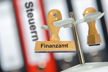 Ein Holzstempel ist beschriftet mit dem Aufdruck Finanzamt vor Aktenordner mit der Aufschrift Steuern