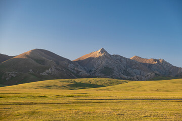 Mountain landscape with green pasture meadow in the foreground and rocky mountains and blue sky in the background shot at  golden hour. Song Kul lake, Naryn Region, Kyrgyzstan, Central Asia.