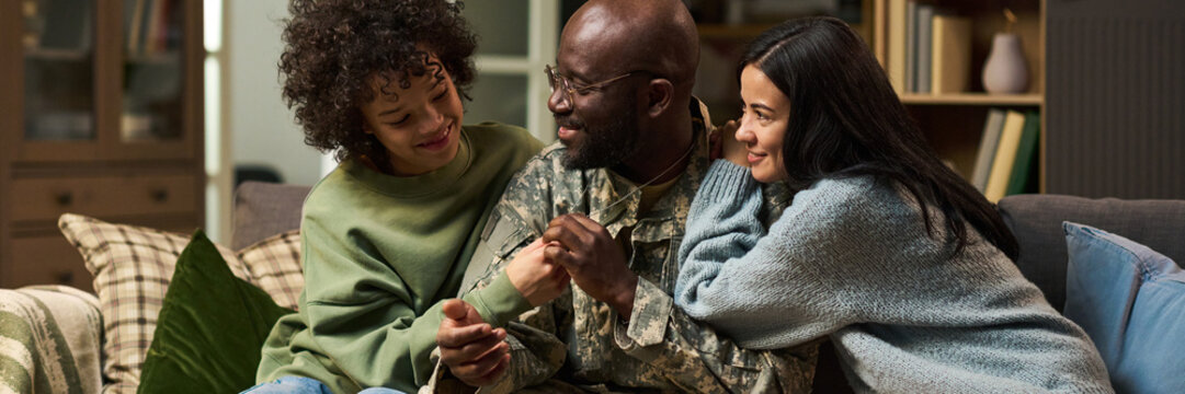 Middle aged Black man in military uniform sitting on couch embracing smiling teenage girl and young adult Caucasian woman, family members showing affection and support at home - Powered by Adobe