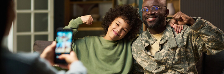 Black middle aged man in military uniform sitting with smiling multiethnic child flexing arms while woman taking photo with smartphone at home, showing family bonding moment