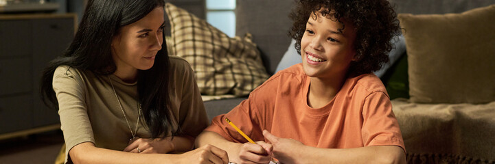 Caucasian woman helping smiling Black teenager with homework at home, both sitting on floor and...