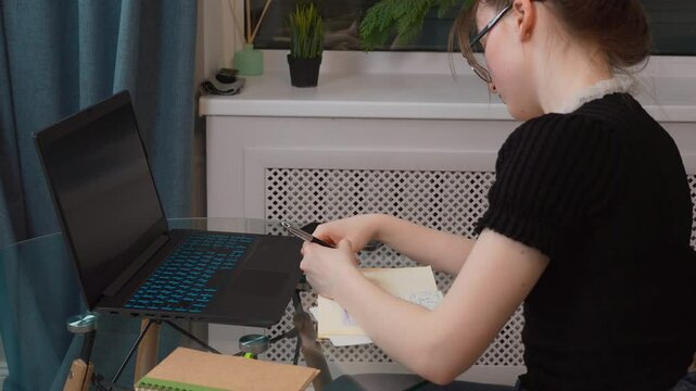 Student Girl Teen With Glasses Writing Notes From Online Lesson, Sitting At Glass Table At Home Workplace, Focusing On Digital Learning With Laptop