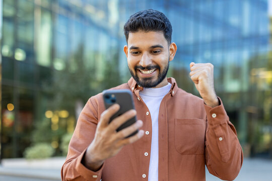 Fototapeta A young Indian man smiles with excitement while looking at his phone, celebrating success in front of a modern building.