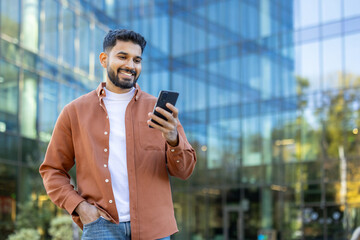 A smiling man of Indian descent looks at his phone while standing outside a modern building during...
