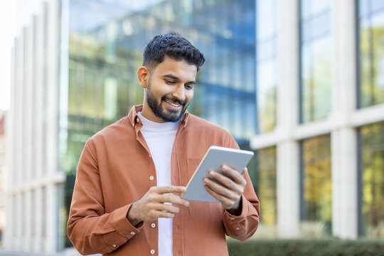 A smiling man with a beard uses a tablet outside a modern building on a sunny day, looking at the screen.