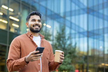 A smiling Indian man holds a coffee cup and smartphone in front of a modern glass building on a...