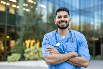 Smiling healthcare professional in scrubs with a stethoscope, standing outdoors in front of a...