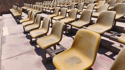 Rows of old, sun-faded plastic stadium seats in an outdoor arena under bright sunlight. The scene...