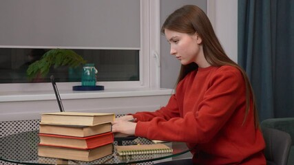 Dedicated student girl teen working on laptop and taking notes in notebook, using stack of books, showing dedication to education and online learning or exam preparation