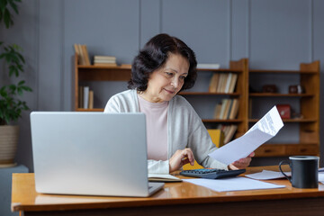 Middle aged senior woman sit with laptop and paper document. Pensive older mature lady reading...