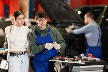 Young woman in a car dealership discusses with a mechanic the replacement of parts and checking the...