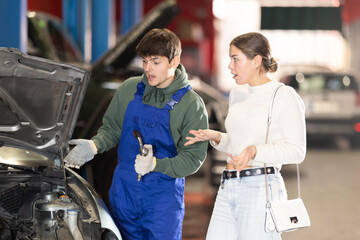 Male mechanic advises young woman client on repairing under hood of car in car service station