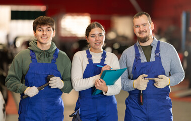 Team of positive auto mechanics, two men and one woman, wearing blue overalls and posing, ready to...