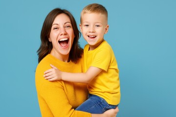 Smiling Mother in yellow sweater holds her happy son in yellow shirt and jeans in her arms. They both look at the viewer, laughing against a light blue background