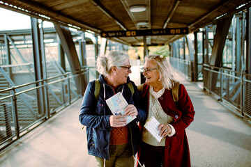 Senior women laughing and holding travel maps at train station platform