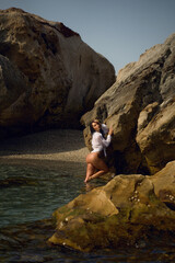 Young brunette woman in a soaked white shirt kneels near a rocky shore, posing seductively under...