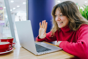 Smiling freelancer waving at a laptop screen during a video call in a cozy cafe, embracing the joys...