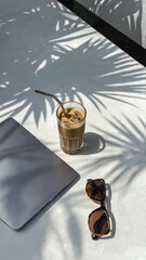 A glass of iced coffee sits on a table with a laptop and sunglasses. Shadows of palm leaves create a tropical atmosphere.