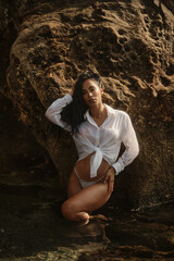 Young brunette woman in a soaked white shirt kneels near a rocky shore, posing seductively under...