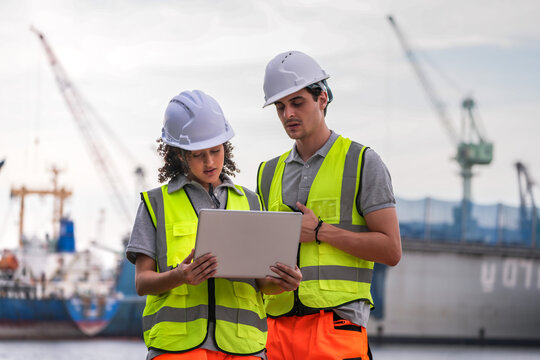 Two engineers wearing safety vests and helmets inspect port operations near the waterfront. They use a laptop and walkie-talkie, symbolizing teamwork, logistics, and marine industry work.