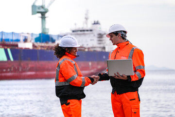 Two port engineers in high-visibility orange uniforms and hard hats communicate while inspecting...