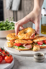 Hand reaches for homemade bagle sandwiches with ham, soft cheese and vegetables on kitchen table background