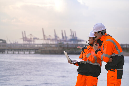 Two engineers wearing safety uniforms and hard hats communicate using a radio while reviewing data on a laptop by the sea during sunset. Maritime industrial setting.