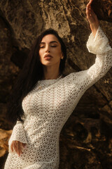 Young brunette model in a white crochet dress poses barefoot on coastal rocks, with one leg raised...