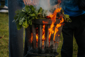 A metal fire basket with tall, bright orange flames burns on a grassy lawn, with embers rising into...